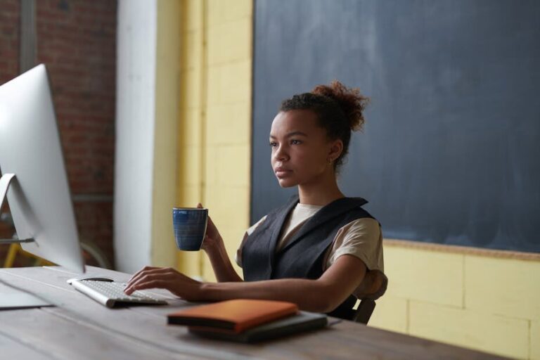 A professional young woman concentrating on work at a desk with a computer and coffee cup in hand.
