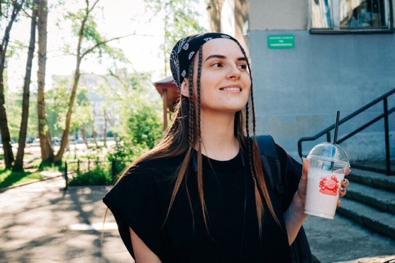 Smiling young woman with a bandana and braids holds a beverage while enjoying a sunny day outside.
