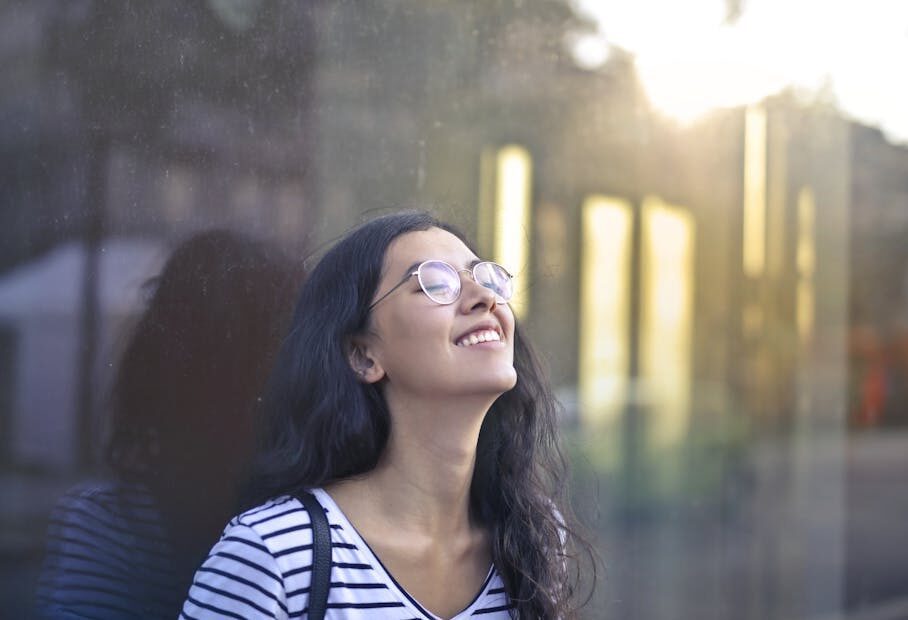A woman with eyeglasses and striped shirt smiling serenely while leaning against a glass wall outdoors.