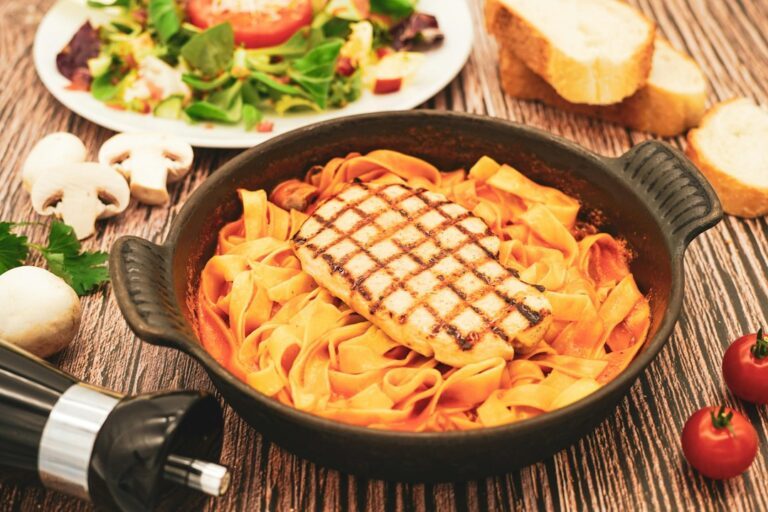 Grilled chicken breast on tagliatelle pasta with creamy tomato sauce, served in a cast iron pan with fresh salad, mushrooms and bread. A classic Italian-Mediterranean restaurant main course — perfect for restaurant menus, food blogs and gastronomy websites.. Imagem ilustrativa para Receita de Macarrão com Frango Marry Me.